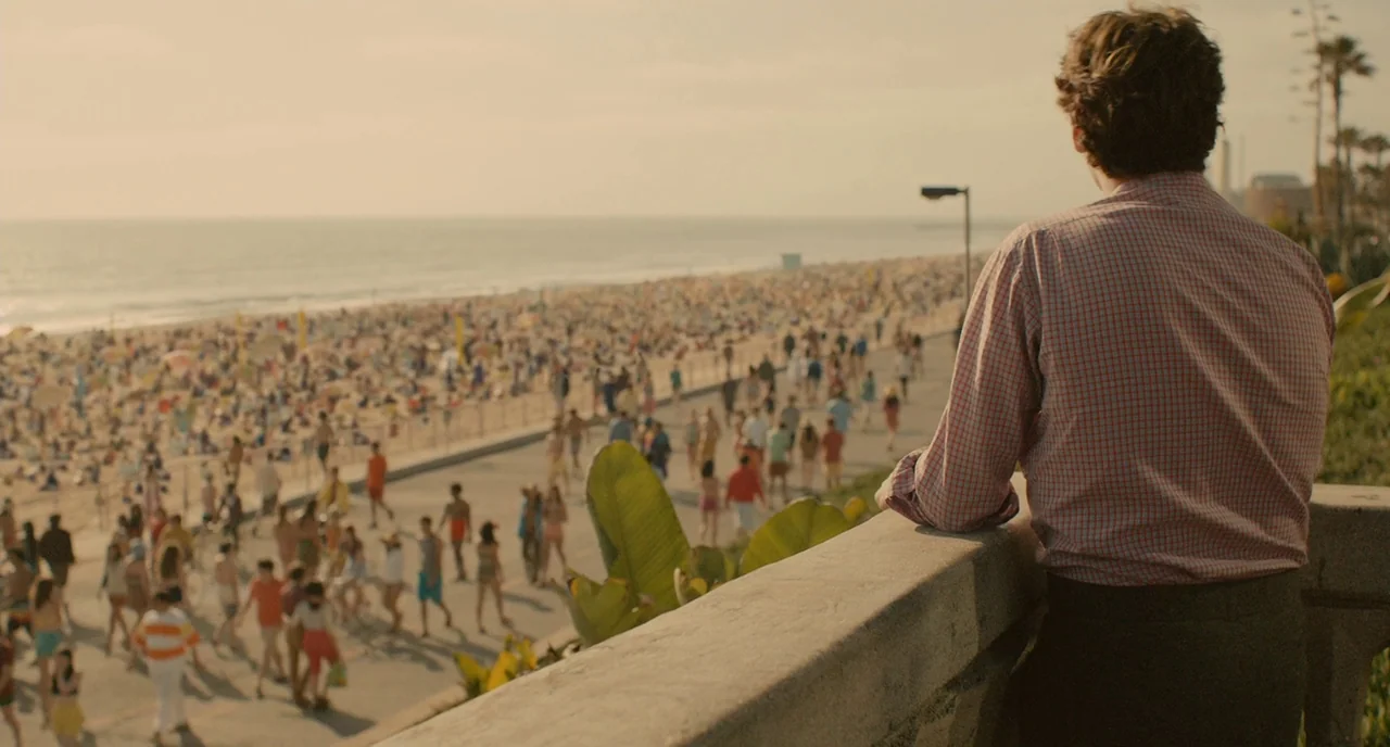 Her (2013) movie still: medium — A sunny coastal boardwalk overlooks a crowded sandy beach filled with many people. A man in a checkered shirt…
