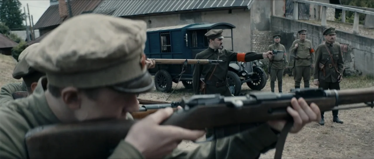 The Rifleman (2019) movie still: medium — A group of soldiers stand in a dirt courtyard next to a vintage dark blue vehicle. One soldier aims a long…