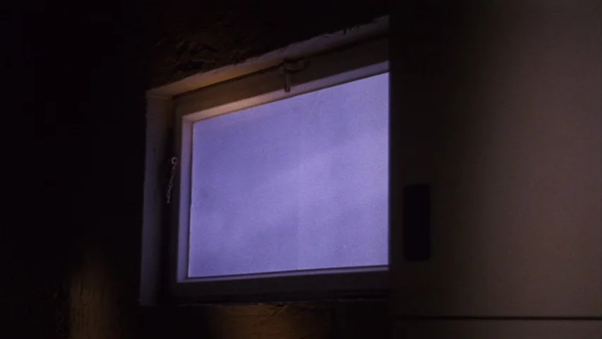Black Sabbath (1963) movie still: closeup — A small rectangular window is set into a dark, textured wall. The frosted pane shows a uniform violet…