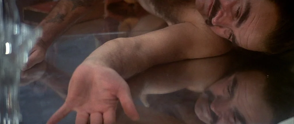 Zardoz (1974) movie still: closeup — A man lies with his face and arm resting on a dark reflective surface. The reflection of his head is visible in…