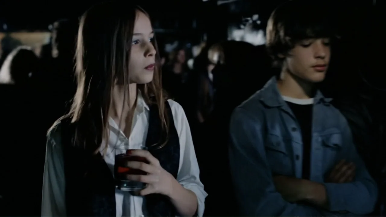 Christiane F. (1981) movie still: medium — A dimly lit indoor gathering with crowded silhouettes visible in the dark background. A young girl stands…