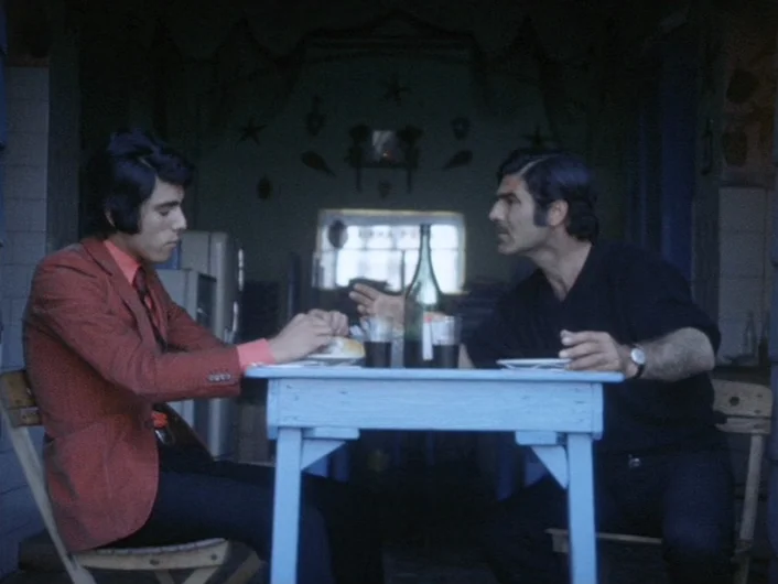 Eika Katappa (1969) movie still: medium — A dimly lit space features a small, pale blue wooden table in the foreground. Two men sit opposite each…