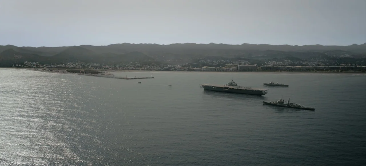 Devotion (2022) movie still: establishing — A wide aerial view shows a coastal bay with hilly terrain in the distance. Several naval vessels…