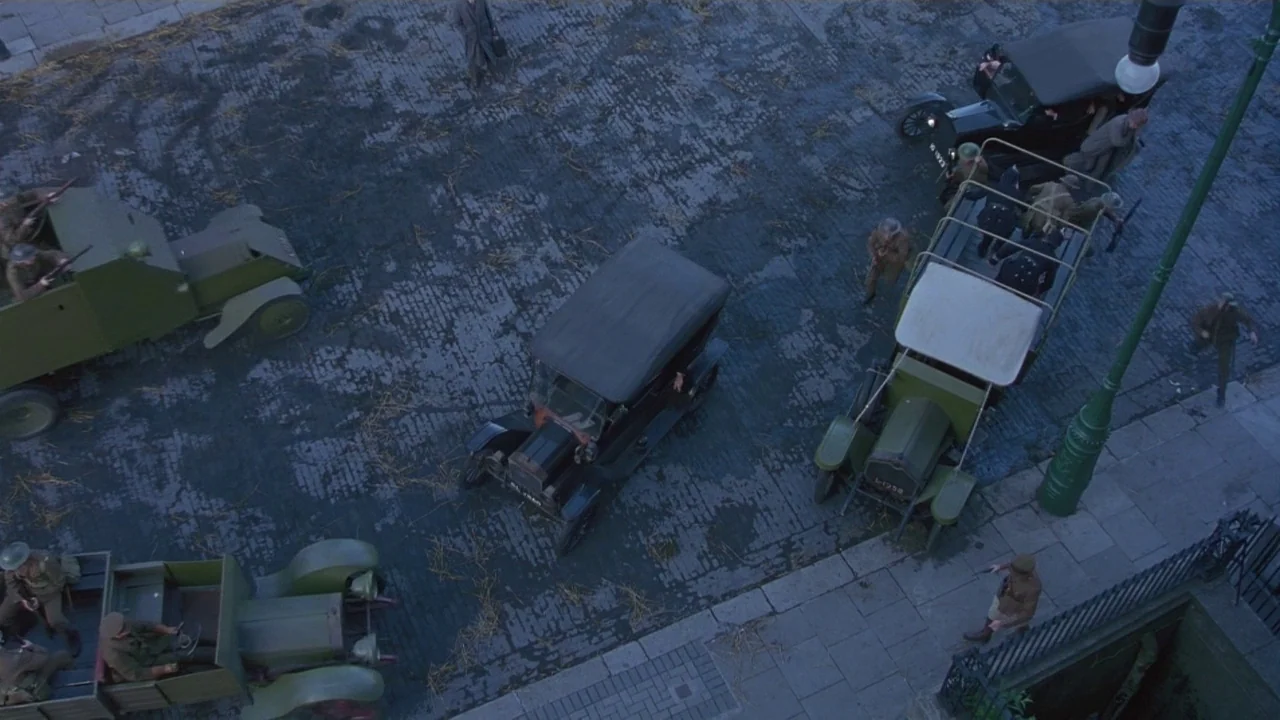 Michael Collins (1996) movie still: establishing — A wet cobblestone city intersection is occupied by early 20th-century military vehicles and…