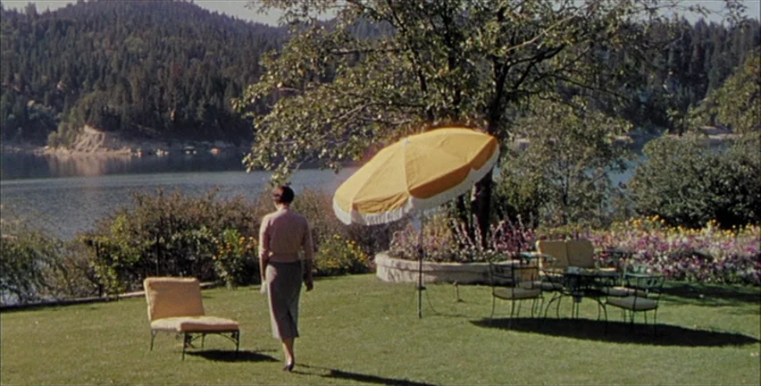 Magnificent Obsession (1954) movie still: wide — A green lawn sits on the edge of a large lake with dense forest mountains. A woman walks across the…