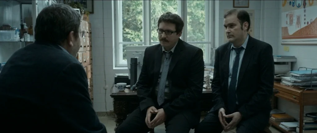 Graduation (2016) movie still: medium — A clinical office contains a large wooden desk and anatomical posters on the walls. Three men in formal…