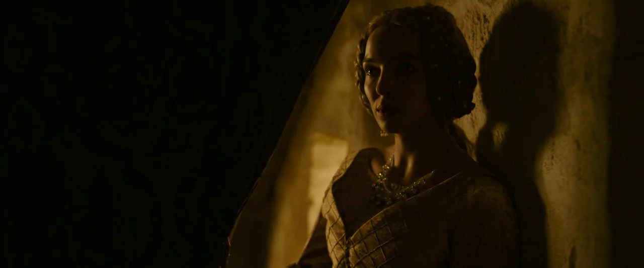 The Last Duel (2021) movie still: medium — A dim, textured stone chamber has a heavy dark curtain hanging in the foreground. A woman with blonde…