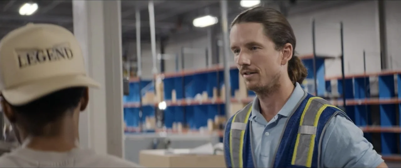 The Forge (2024) movie still: medium — A warehouse interior features tall metal shelving units painted blue in the background. A man in a safety…