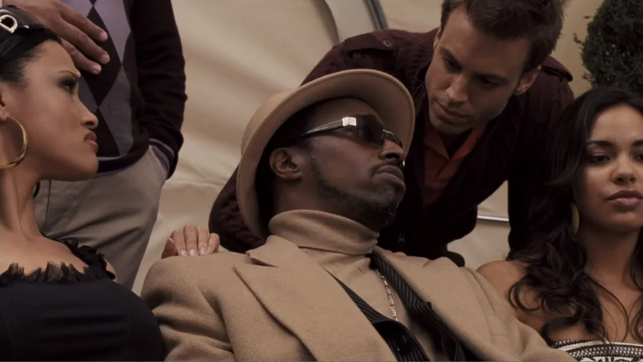 Redline (2007) movie still: medium — An outdoor patio area features a small group of four people together. A man in a fedora sits centrally while…