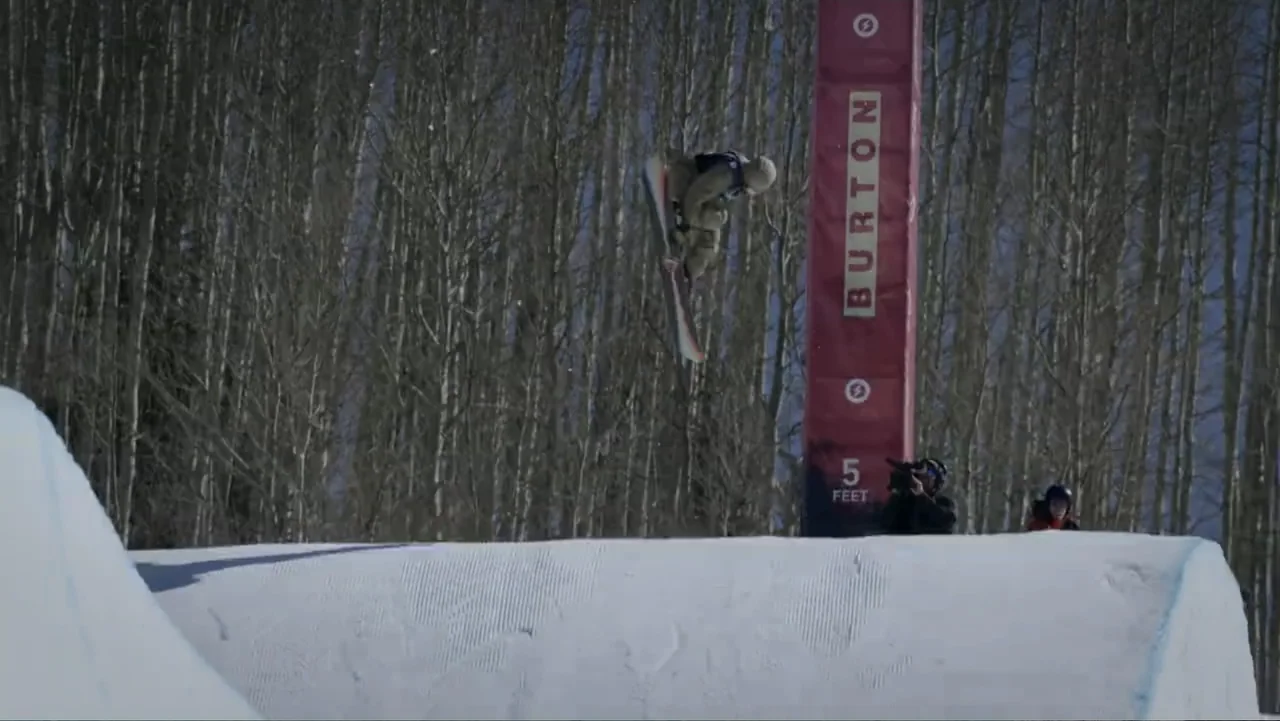 Dear Rider: The Jake Burton Story (2021) movie still: wide — A snowy ski terrain park features a red vertical safety banner and forest backdrop. A…