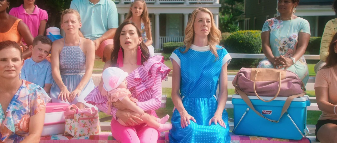 Greener Grass (2019) movie still: medium — A group of women and children sit on metal bleachers outdoors in front of a house. The group appears to…