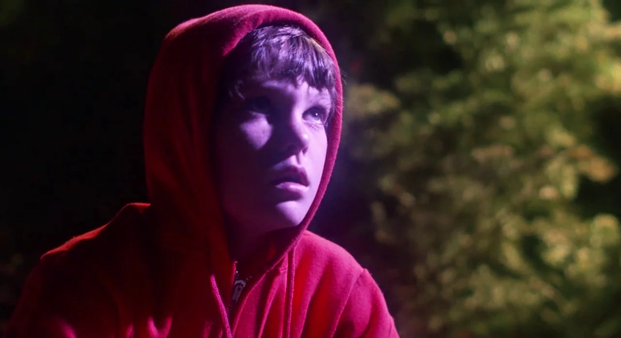 E.T. The Extra-Terrestrial (1982) movie still: closeup — A dark night setting featuring blurred trees in the background. A young boy in a red hoodie…