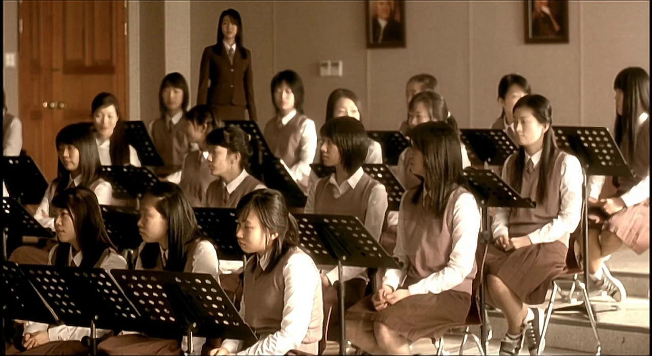 Voice (2005) movie still: wide — A school music room with rows of seated students holding music stands. The room features beige walls adorned with…