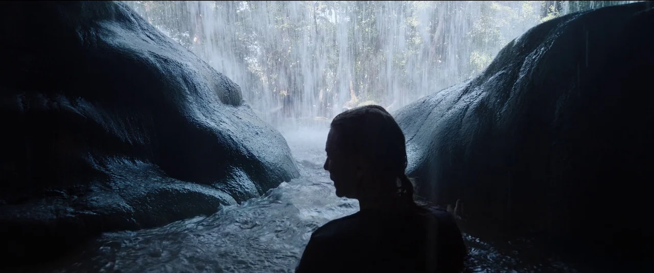 Tár (2022) movie still: medium — A dark cave interior frames a view of a waterfall cascading down outside. A young woman stands waist-deep in the…