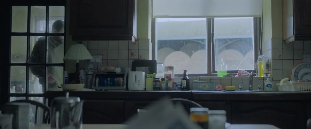 Bring Them Down (2025) movie still: wide — A cluttered kitchen with tiled backsplashes and a window looking out onto exterior greenery. A man stands…
