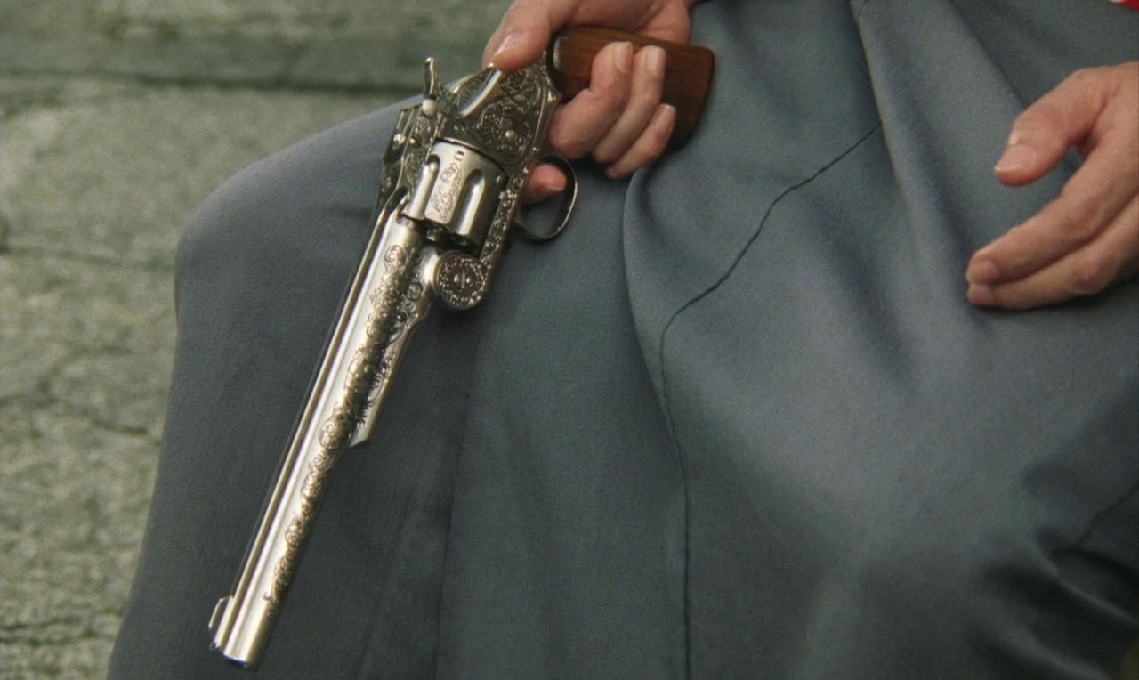 Yumeji (1991) movie still: closeup — A close-up view of a highly detailed, engraved silver revolver held on a lap. The person is wearing gray fabric…