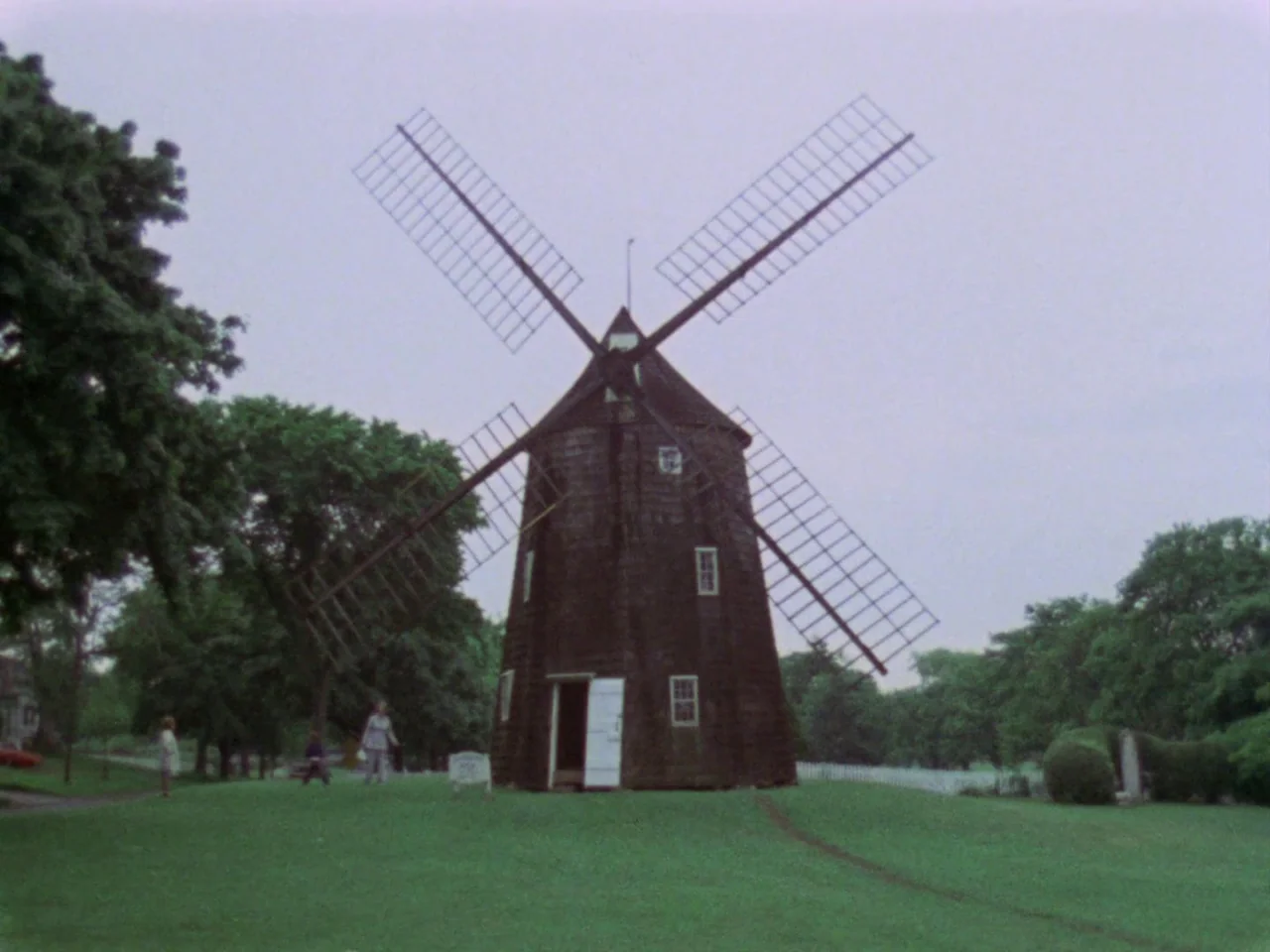 Grey Gardens (1975) movie still: wide — A dark wooden windmill with large sails stands on a green, grassy slope. Three small human figures walk in…
