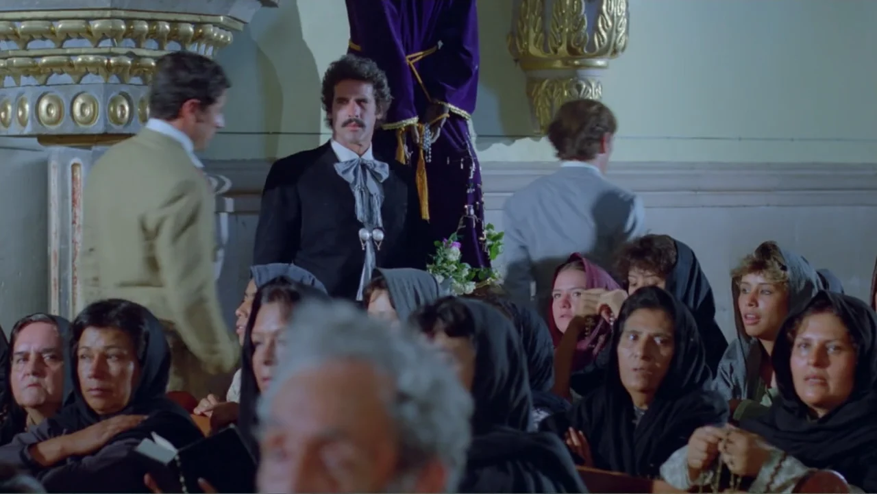 Toña Machetes (1985) movie still: medium — A church interior features wooden pews filled with women wearing black head coverings. A man in a suit…