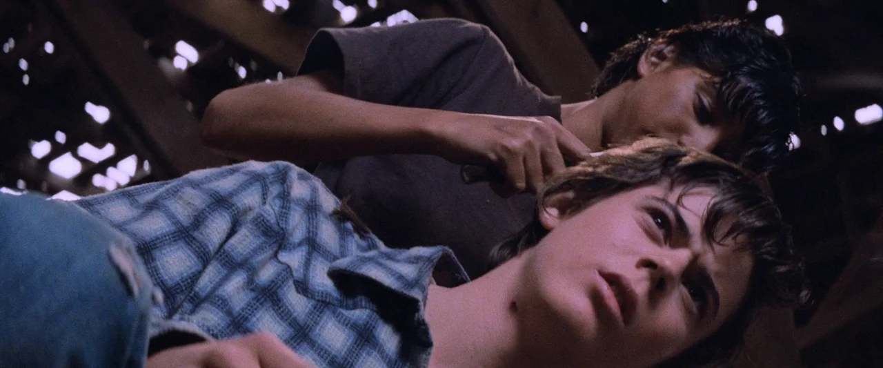 The Outsiders (1983) movie still: medium — A dimly lit wooden interior with light leaking through small holes in the walls. One person uses scissors…