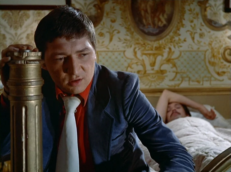 Fox and His Friends (1975) movie still: medium — A man in a blue blazer leans against a golden ornate bedpost. A person with a mustache lies in the…