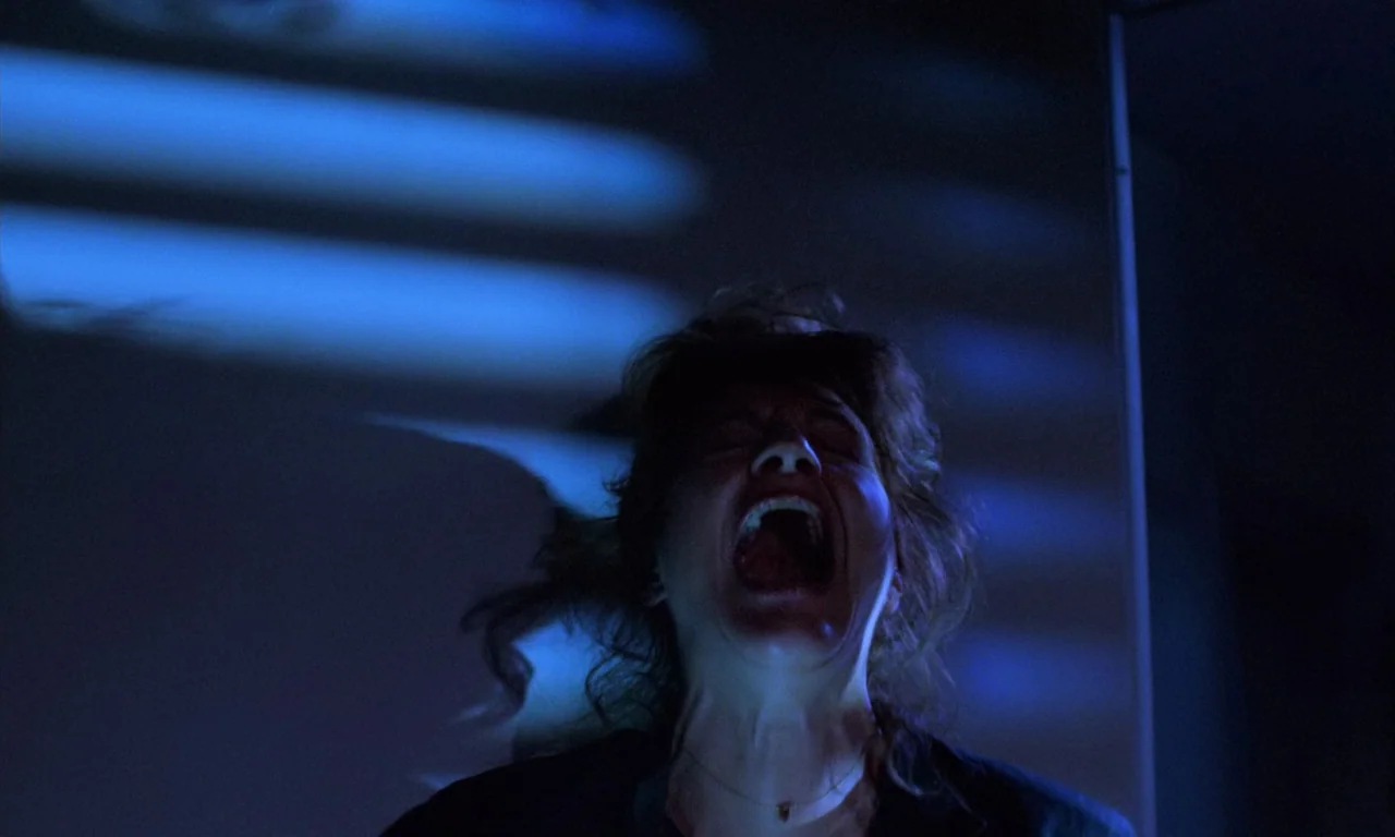 Demons 2 (1986) movie still: closeup — A dimly lit interior space is bathed in horizontal bands of blue light. A woman is in the foreground, head…