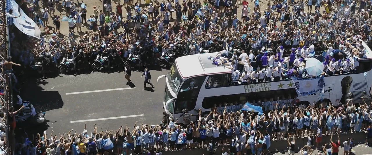 I Choose to Believe (2023) movie still: wide — A sunny asphalt highway is packed with a dense crowd of people. A white double-decker bus carries…