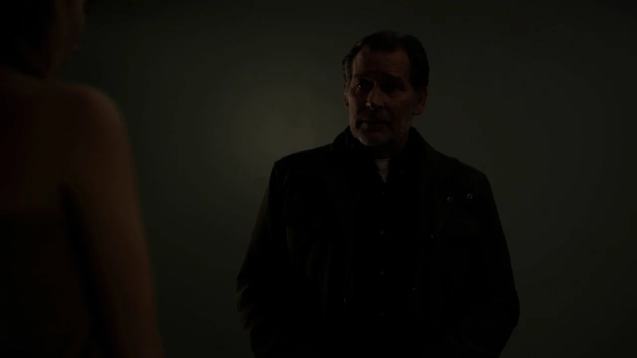 The Blackcoat’s Daughter (2015) movie still: medium — A dimly lit, featureless space frames an older man in a utility jacket. The man faces an…