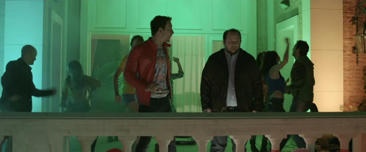 Perdida (2018) movie still: fullbody — A stone balcony overlooks a brightly lit interior party room tinted with green light. Two men stand in the…