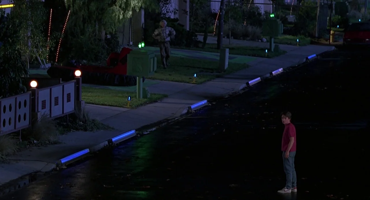 Back to the Future II (1989) movie still: wide — A dark residential suburban street at night features glowing blue curb lights and futuristic…