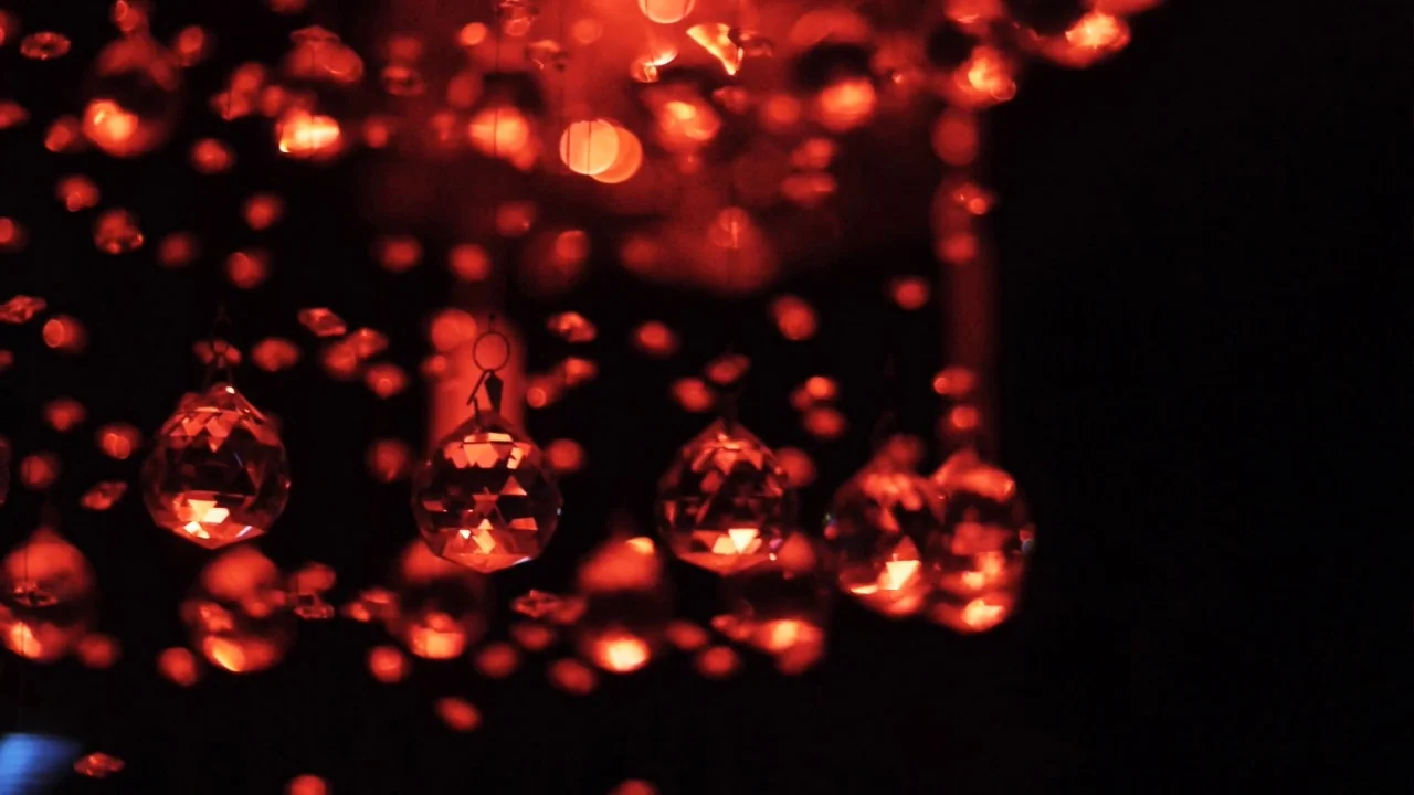 Counting (2015) movie still: closeup — A close-up view of a chandelier with multiple hanging crystal prisms. Red light glows intensely within the…