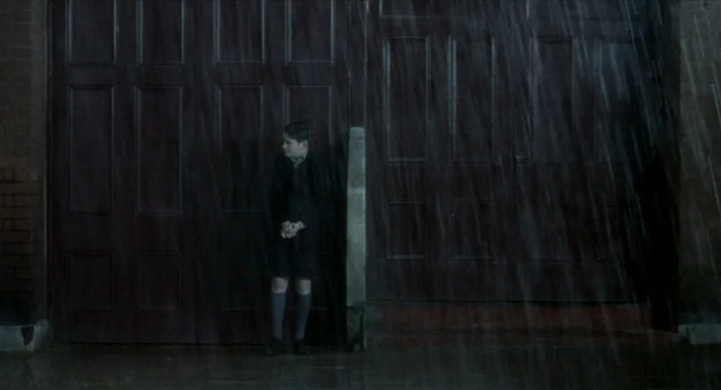 The Long Day Closes (1992) movie still: fullbody — A small child stands on a sidewalk in the dark during a rainstorm. The child is positioned before…