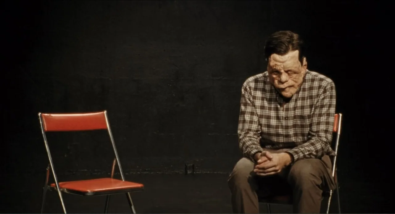A Different Man (2024) movie still: medium — A stark dark void features two metal folding chairs placed on a flat floor. A man with facial scarring…
