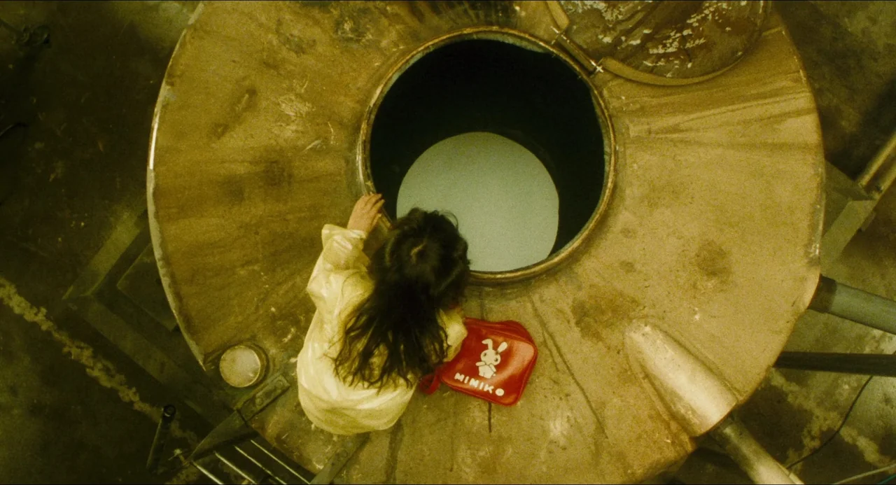 Dark Water (2002) movie still: wide — A top-down view shows a child standing on a large metallic industrial machine. She looks into an open circular…