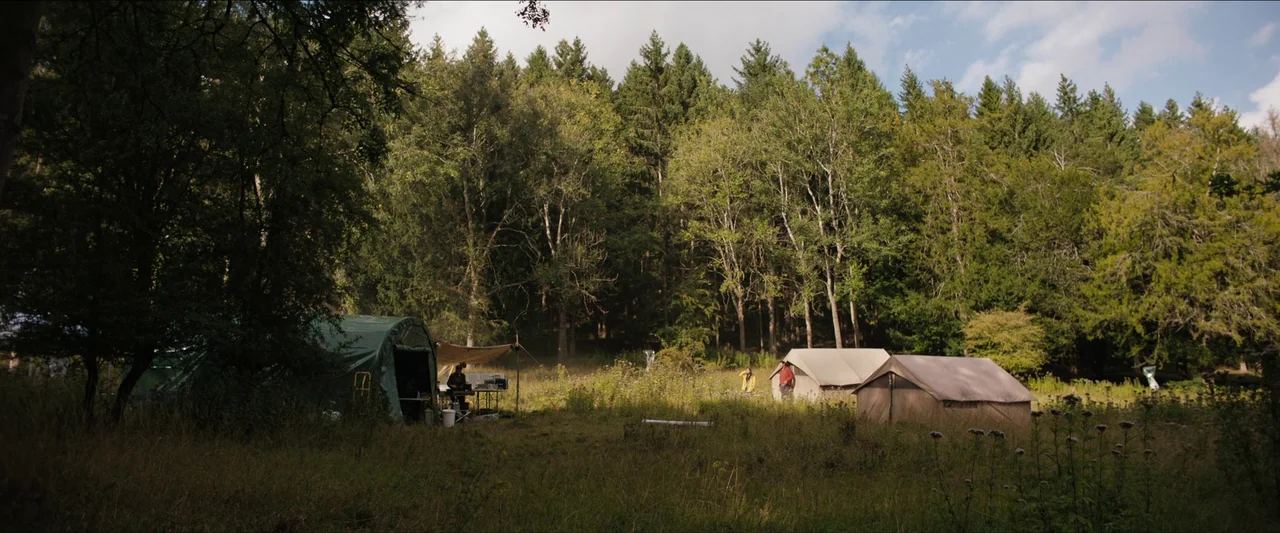 In The Earth (2021) movie still: wide — A grassy clearing at the edge of a dense forest contains several camping tents. Three people stand in the…