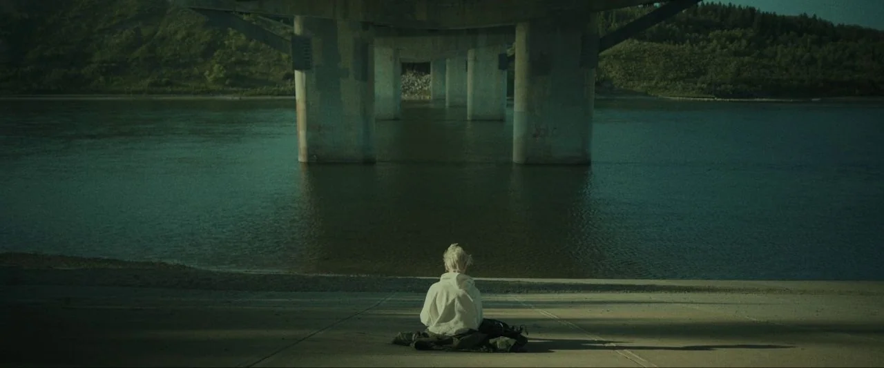 Come True (2020) movie still: wide — A wide shot shows a person sitting on a concrete ledge beneath a massive bridge. A river flows in the…