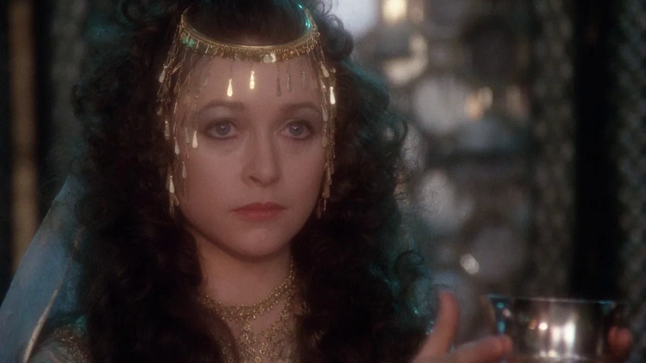 Excalibur (1981) movie still: closeup — A dimly lit fantasy interior features a woman adorned in intricate gold jewelry. She looks forward while…