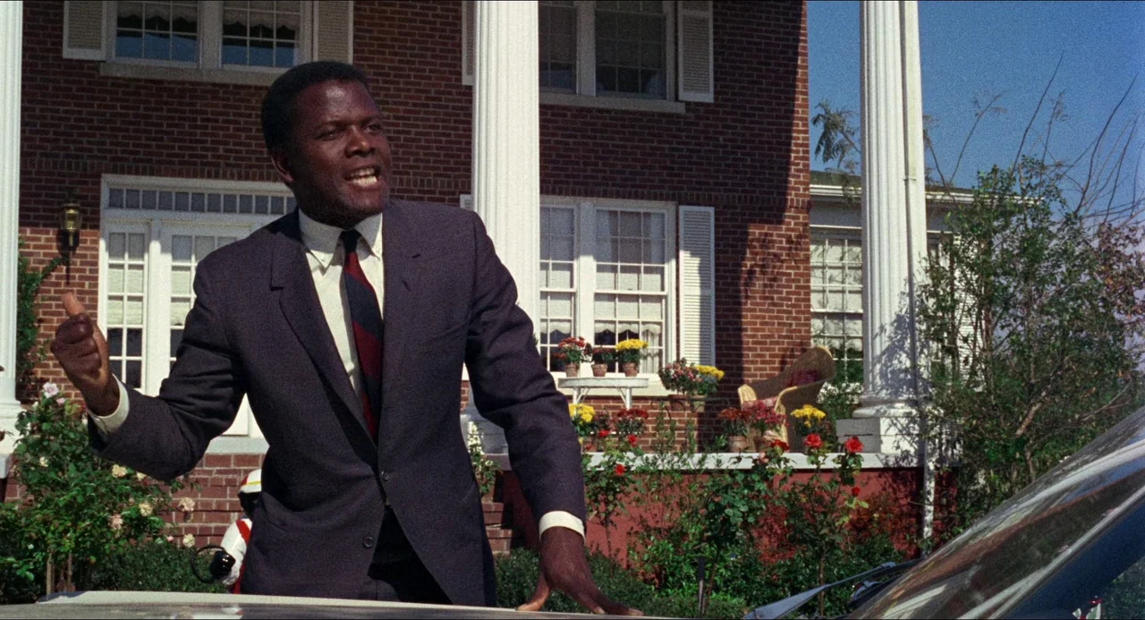 In the Heat of the Night (1967) movie still: medium — A classic brick colonial house with white columns stands behind a manicured suburban lawn. A…