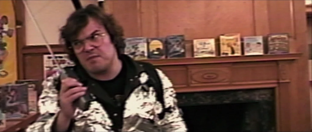 Be Kind Rewind (2008) movie still: medium — A man stands in front of a wooden mantle with several books displayed. The individual is holding a…