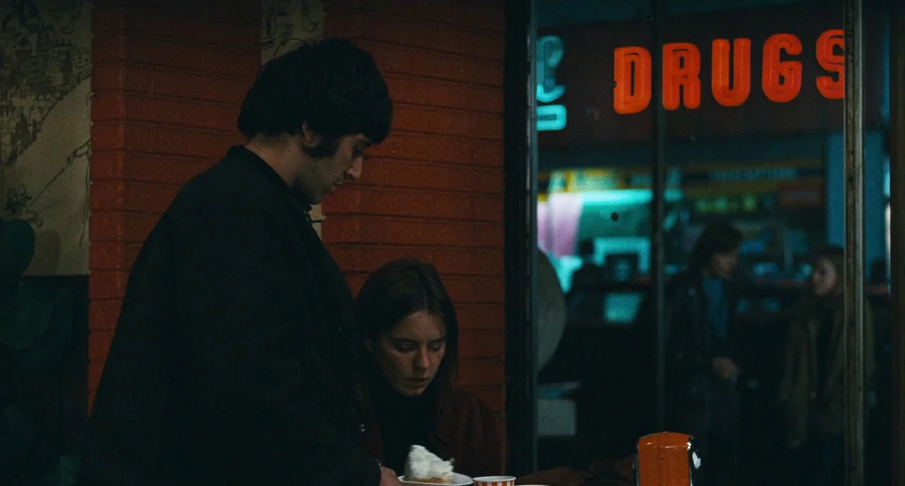 The Panic in Needle Park (1971) movie still: medium — A dark diner booth sits next to a glass window looking out at night. A man stands beside a…