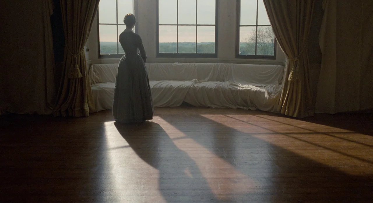 Jane Eyre (2011) movie still: wide — An empty drawing room features draped furniture and polished dark wood floorboards. A woman stands with her…