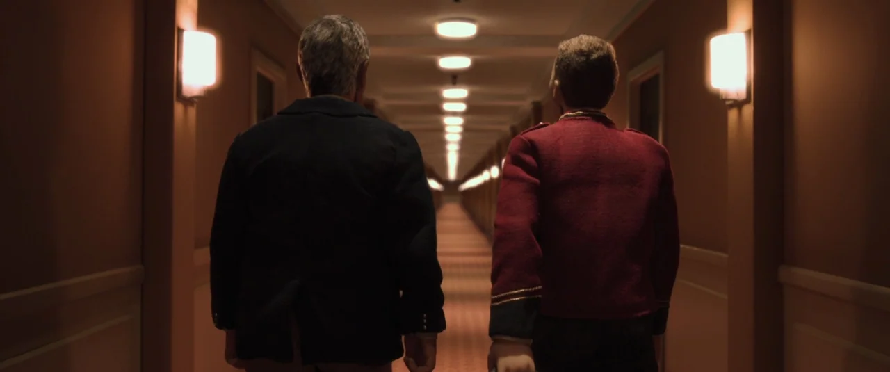 Anomalisa (2015) movie still: fullbody — An extremely long, symmetrical indoor corridor has patterned carpeting and repeating wall sconces. Two men…