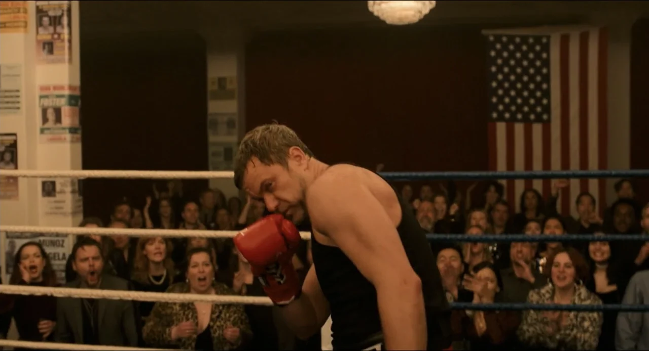 Karate Kid: Legends (2025) movie still: medium — A boxing ring filled with a cheering crowd under bright overhead lights. A male fighter in a black…