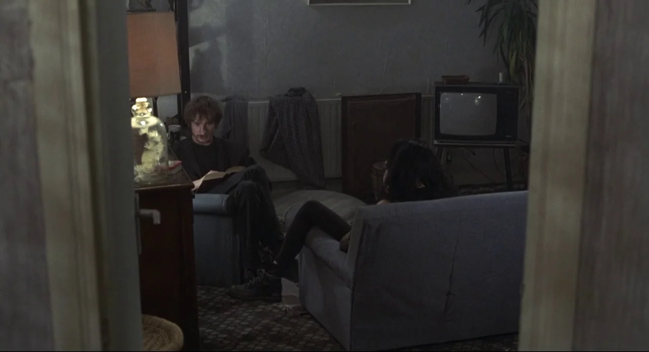 Naked (1993) movie still: wide — A sparsely furnished room contains two armchairs, a television set, and a potted plant. A man reads a book in one…