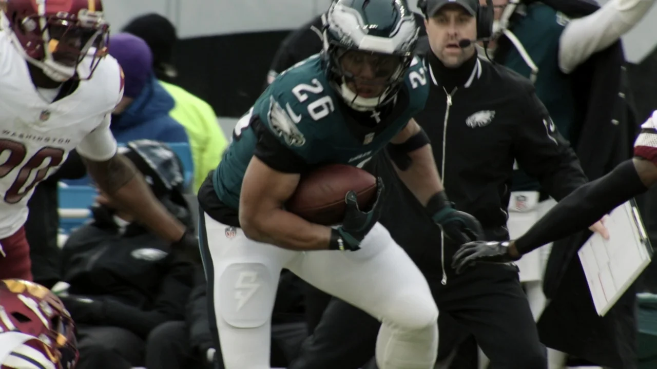 SAQUON (2025) movie still: medium — A professional football player runs with a ball on a green turf field. Other players and coaching staff are…
