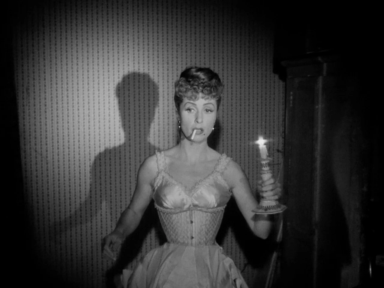 Le Plaisir (1952) movie still: medium — A dimly lit indoor space features textured wallpaper with a repeating diamond pattern. A woman in a corset…