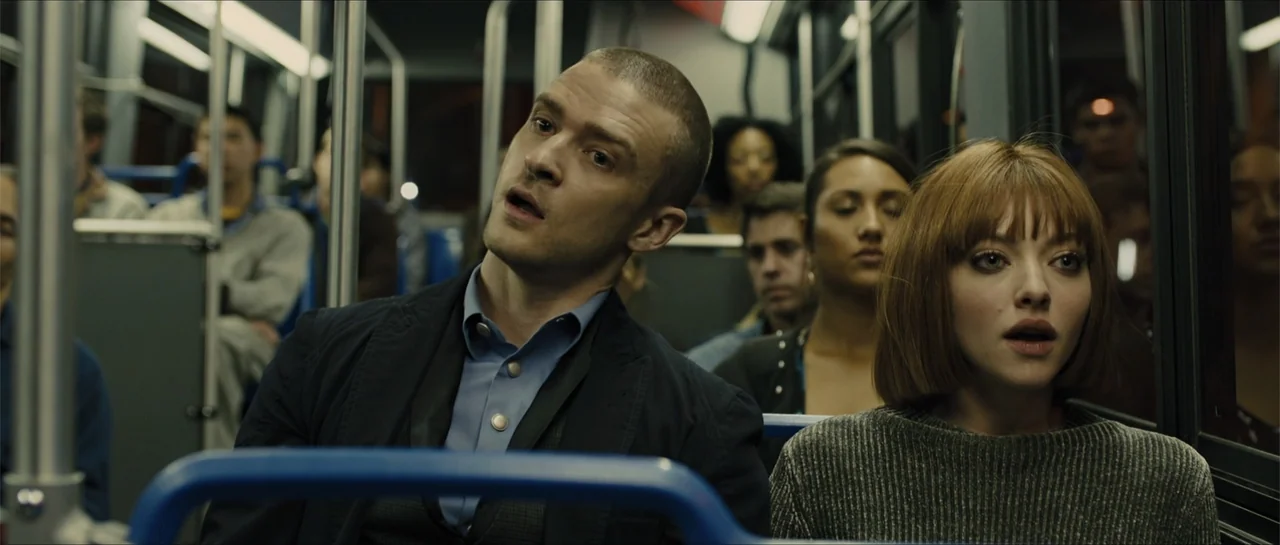 In Time (2011) movie still: medium — Rows of passengers occupy seats on a crowded modern bus at night. A man and a woman in the foreground gaze…