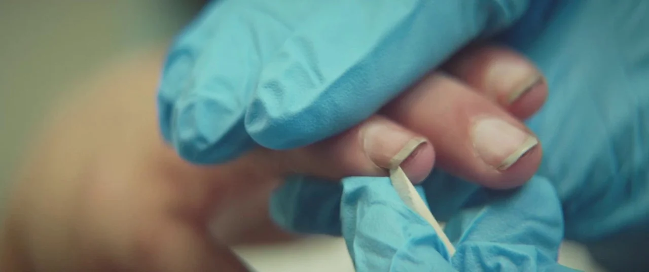 The Strange Ones (2017) movie still: An extreme close-up capturing a moment of precise manual care, where blue-gloved hands perform a detailed task…