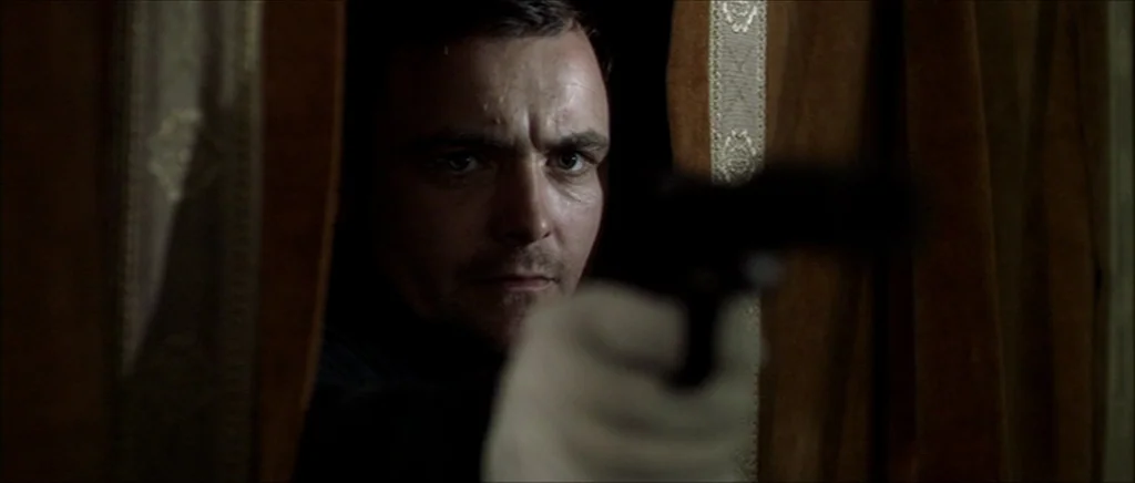Kill List movie still: closeup — A man peers through a gap between heavy brown curtains in a dimly lit room. He aims a handgun forward while wearing…
