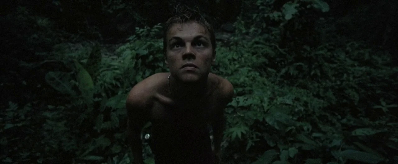 The Beach (2000) movie still: medium — A dense, dark tropical jungle environment with thick vegetation and large fern leaves. A young man stands…