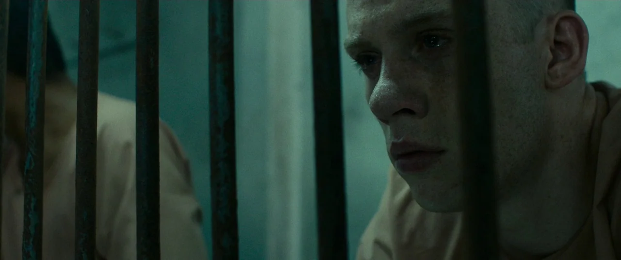 A Prayer Before Dawn (2017) movie still: closeup — A dark, cramped space defined by vertical rusted metal bars separating the foreground. A young…